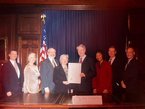 Group of business professionals posing for a formal photo in an ornate room and holding a proclamation document with a gold seal.