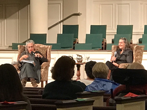 Two women hold microphones and sit in plush chairs on a stage during a crowded author event.